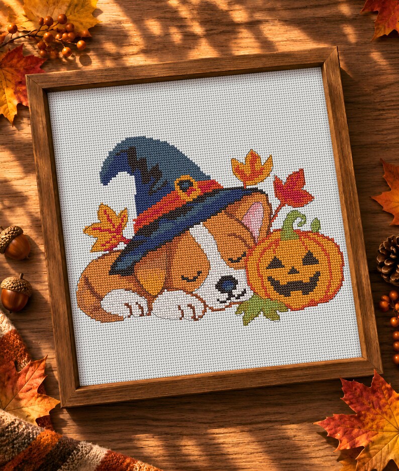 May include: A cross-stitch embroidery in a wooden frame featuring a sleeping puppy wearing a blue witch hat with an orange band. The puppy is positioned next to a carved orange jack-o'-lantern, surrounded by several small, colourful autumn leaves.
