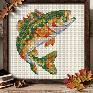 May include: A framed cross-stitch embroidery featuring a leaping fish rendered in a vibrant palette of green, orange, yellow, and blue threads. The design is set against a white fabric background, enclosed within a dark brown wooden frame.