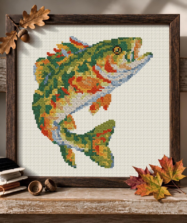 May include: A framed cross-stitch embroidery featuring a leaping fish rendered in a vibrant palette of green, orange, yellow, and blue threads. The design is set against a white fabric background, enclosed within a dark brown wooden frame.