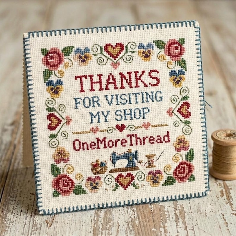May include: A square cross-stitch sampler on cream fabric with a dark blue blanket-stitched border. It features floral motifs, hearts, and the text: THANKS FOR VISITING MY SHOP OneMoreThread, with a small sewing machine and thread spool.