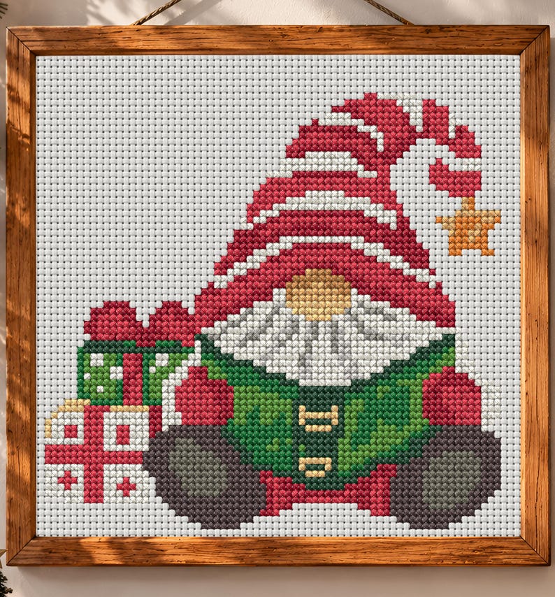 May include: A cross-stitch pattern in a wooden frame featuring a gnome wearing a large red hat with a yellow star at the tip. The gnome has a white beard, a green tunic, and dark gray boots, and is positioned next to two wrapped gift boxes.