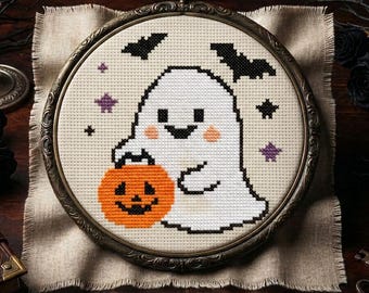 May include: A cross-stitch embroidery in a round, ornate bronze-colored frame featuring a white ghost with a smiling face, pink cheeks, and black eyes holding an orange jack-o'-lantern, surrounded by black bats and small purple stars.