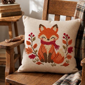 May include: A square throw pillow with a beige fabric base featuring a cross-stitch design of a fox wearing a red scarf, surrounded by autumn leaves and berries in shades of orange, red, and brown, centered on the front of the pillow.