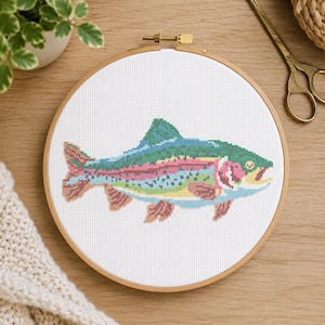 May include: A circular wooden embroidery hoop containing a cross-stitch pattern of a rainbow trout. The fish features a green back, a pink lateral stripe, and blue and yellow accents on a white fabric background with a visible grid structure.