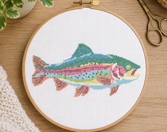 May include: A circular wooden embroidery hoop containing a cross-stitch pattern of a rainbow trout. The fish features a green back, a pink lateral stripe, and blue and yellow accents on a white fabric background with a visible grid structure.