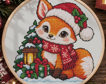 May include: A cross-stitch embroidery in a wooden hoop featuring a cartoon fox wearing a red Santa hat and a red scarf with white snowflakes. The fox sits next to a green gift box, a lit lantern, and a small pine tree with red berries.