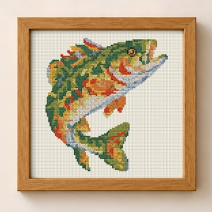 May include: A square wooden frame containing a cross-stitch embroidery of a fish. The fish is depicted in a leaping position with a color palette of green, orange, yellow, and blue threads against a white fabric background with a visible grid.