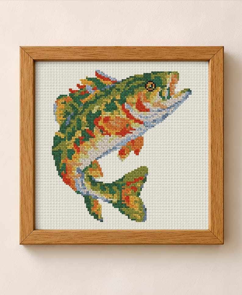 May include: A square wooden frame containing a cross-stitch embroidery of a fish. The fish is depicted in a leaping position with a color palette of green, orange, yellow, and blue threads against a white fabric background with a visible grid.