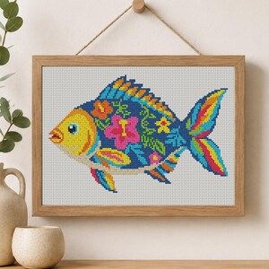 Pode incluir: Um bordado em ponto cruz de um peixe numa moldura de madeira clara. O peixe tem a cabe&ccedil;a amarela, o corpo azul-escuro com flores de hibisco cor-de-rosa e laranja, e uma cauda multicolorida com riscas azuis, amarelas e cor-de-rosa.