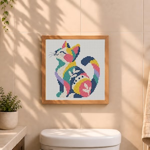 May include: A square cross-stitch embroidery featuring a stylized cat in a seated position. The cat is composed of vibrant, multi-colored geometric patterns including pink, yellow, blue, and green, all set against a white grid fabric in a wooden frame.