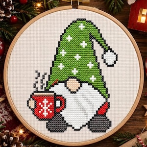 May include: A cross-stitch embroidery in a round wooden hoop featuring a gnome with a green hat decorated with white plus signs, a large white beard, and dark grey boots. The gnome holds a red mug with a white snowflake design and steam.