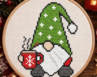 May include: A cross-stitch embroidery in a round wooden hoop featuring a gnome with a green hat decorated with white plus signs, a large white beard, and dark gray boots. The gnome holds a red mug with a white snowflake design and steam.