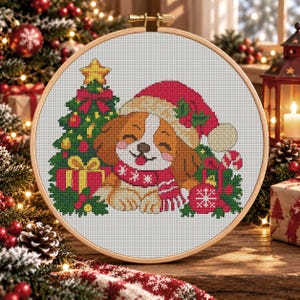 May include: A circular wooden embroidery hoop containing a cross-stitch pattern of a brown and white puppy wearing a red Santa hat and a red scarf with white snowflakes. The puppy sits beside a green Christmas tree, gifts, and ornaments.