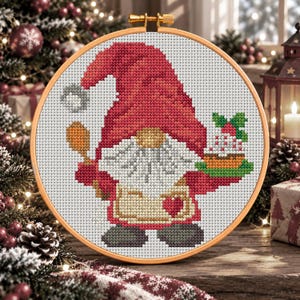 May include: A cross-stitch embroidery in a round wooden hoop featuring a gnome with a large red hat, a white beard, and a tan apron with a red heart. The gnome holds a wooden spoon in one hand and a small cake with green leaves in the other.