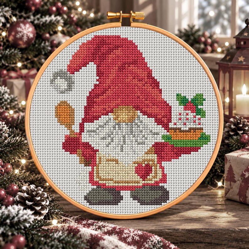 May include: A cross-stitch embroidery in a round wooden hoop featuring a gnome with a large red hat, a white beard, and a tan apron with a red heart. The gnome holds a wooden spoon in one hand and a small cake with green leaves in the other.