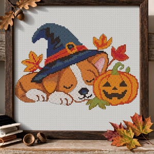 May include: A cross-stitch embroidery in a dark wooden frame depicting a sleeping puppy wearing a blue witch hat with an orange band. Beside the puppy is a carved orange jack-o'-lantern pumpkin with a green stem, surrounded by colourful autumn leaves.