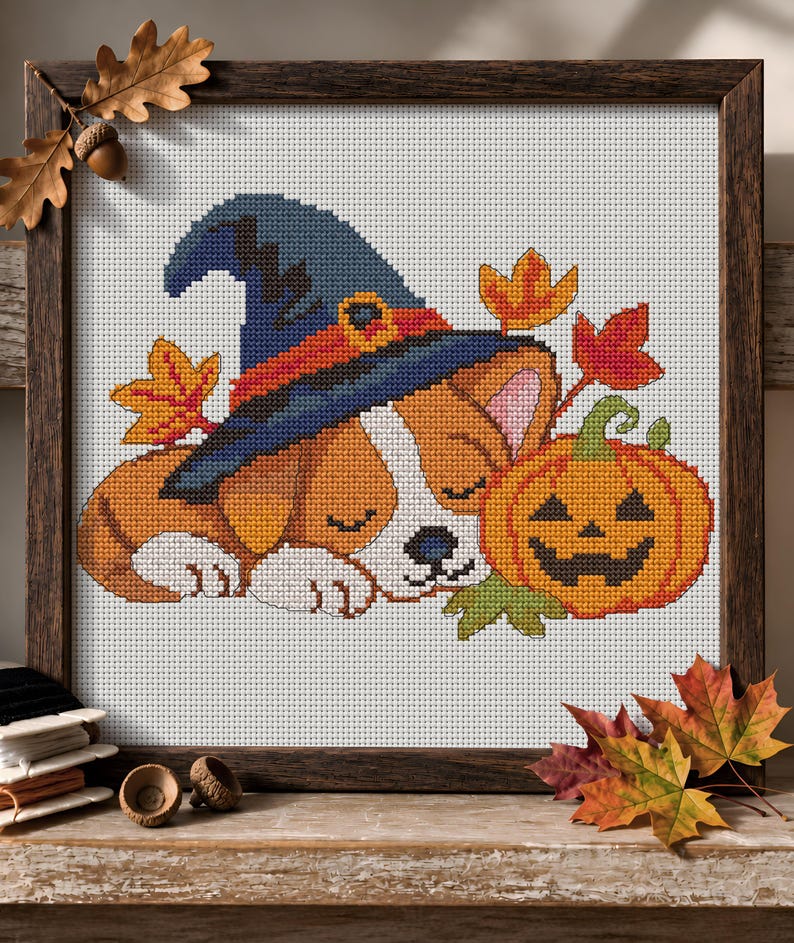 May include: A cross-stitch embroidery in a dark wooden frame depicting a sleeping puppy wearing a blue witch hat with an orange band. Beside the puppy is a carved orange jack-o'-lantern pumpkin with a green stem, surrounded by colourful autumn leaves.