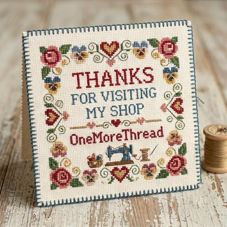 May include: A square cross-stitch sampler on cream fabric with a dark blue stitched border. It features red and blue text reading "THANKS FOR VISITING MY SHOP" and "OneMoreThread" surrounded by a floral border of roses, pansies, and hearts.