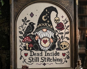 May include: A cross-stitch embroidery in a dark wooden frame featuring a gnome wearing a black wizard hat with a crescent moon, holding a hoop with a heart. The design includes a black bird, red mushrooms, a skull, roses, and the text Dead Inside Still Stitching.