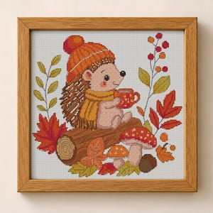 May include: A cross-stitch embroidery in a light wood frame depicting a hedgehog wearing an orange knit hat and yellow scarf, holding a red mug. The scene includes a brown log, red and yellow autumn leaves, two mushrooms, and red berries.