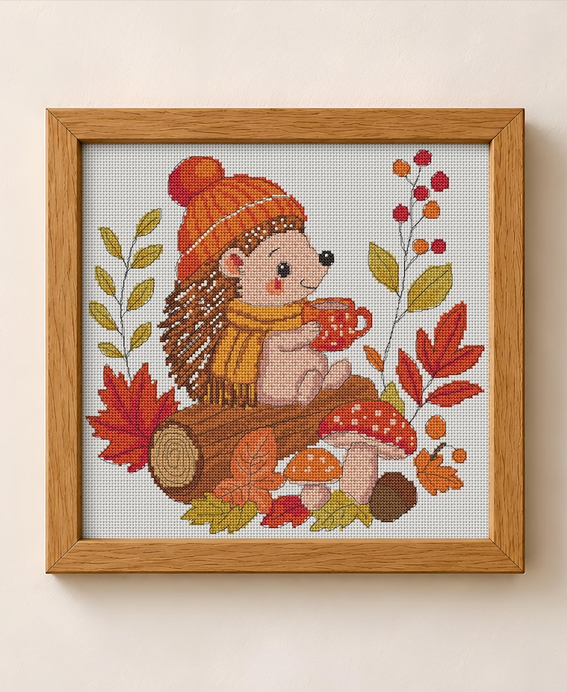 May include: A cross-stitch embroidery in a light wood frame depicting a hedgehog wearing an orange knit hat and yellow scarf, holding a red mug. The scene includes a brown log, red and yellow autumn leaves, two mushrooms, and red berries.