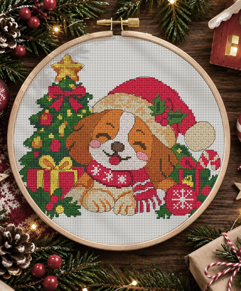 May include: A cross-stitch embroidery in a round wooden hoop featuring a brown and white puppy wearing a red Santa hat and a red scarf. The puppy is surrounded by a decorated Christmas tree, wrapped gifts, holly leaves, and a red ornament.