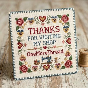 May include: A square cross-stitch sampler on cream fabric with a dark blue stitched border. It features the text "THANKS FOR VISITING MY SHOP" and "OneMoreThread" in red and blue, surrounded by floral motifs, hearts, and a sewing machine.