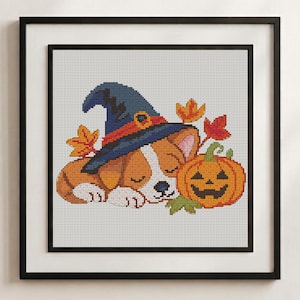 May include: A cross-stitch pattern featuring a sleeping puppy wearing a blue witch hat with an orange band, positioned next to a carved orange jack-o'-lantern and several colourful autumn leaves, all framed in a black square frame.
