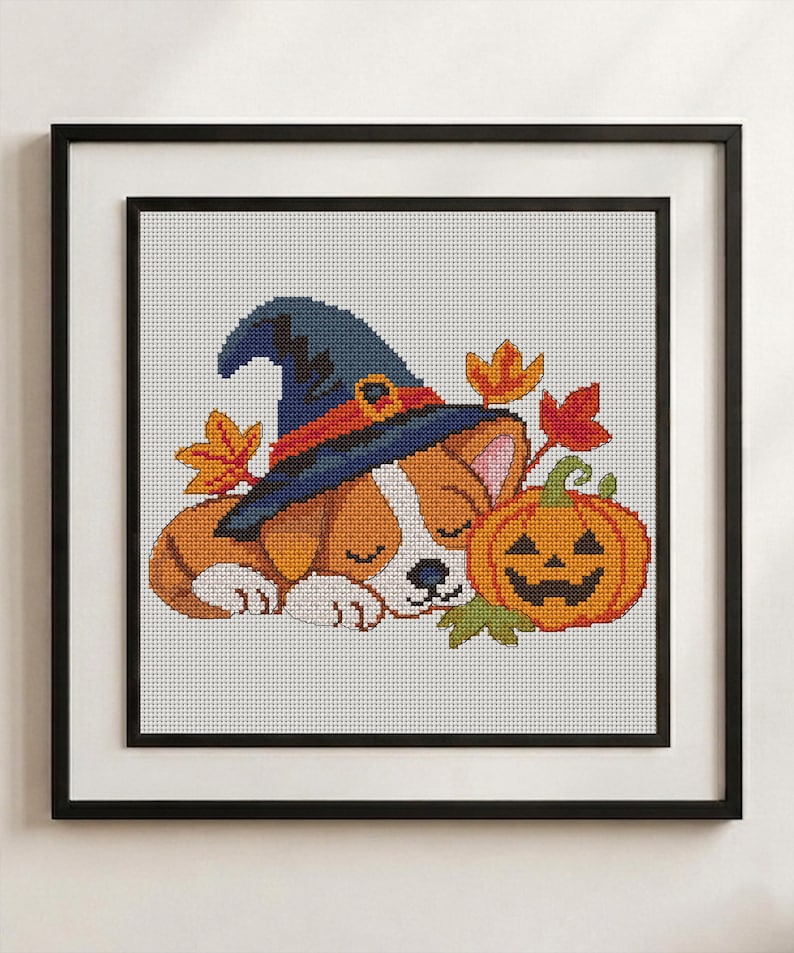 May include: A cross-stitch pattern featuring a sleeping puppy wearing a blue witch hat with an orange band, positioned next to a carved orange jack-o'-lantern and several colourful autumn leaves, all framed in a black square frame.