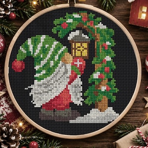 May include: A circular wooden embroidery hoop containing a cross-stitch pattern on black fabric. The design features a gnome wearing a green and white striped hat, a long white beard, and red clothing, standing beside a green Christmas tree.