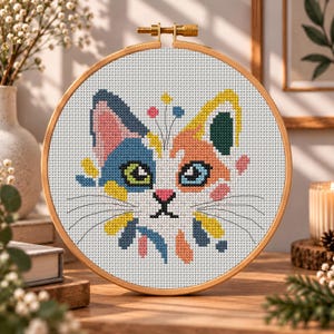 K&ouml;nnte beinhalten: Ein runder Stickrahmen aus Holz mit einem Kreuzstichmuster eines Katzengesichts. Die Katze hat ein mehrfarbiges Design mit blauen, orangefarbenen, gelben und rosafarbenen Flecken auf wei&szlig;em Stoff sowie aufgestickten schwarzen Schnurrhaaren.