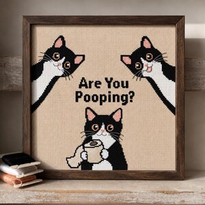 May include: A framed cross-stitch embroidery featuring three black and white tuxedo cats on a beige fabric background. The text "Are You Pooping?" is centered in black letters, with one cat holding a roll of toilet paper in the lower section.