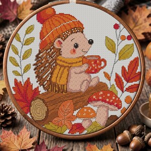 May include: A circular wooden embroidery hoop containing a cross-stitch pattern of a hedgehog wearing an orange knit hat and a yellow scarf, holding a red mug. The scene includes a brown log, red and orange autumn leaves, and two mushrooms.