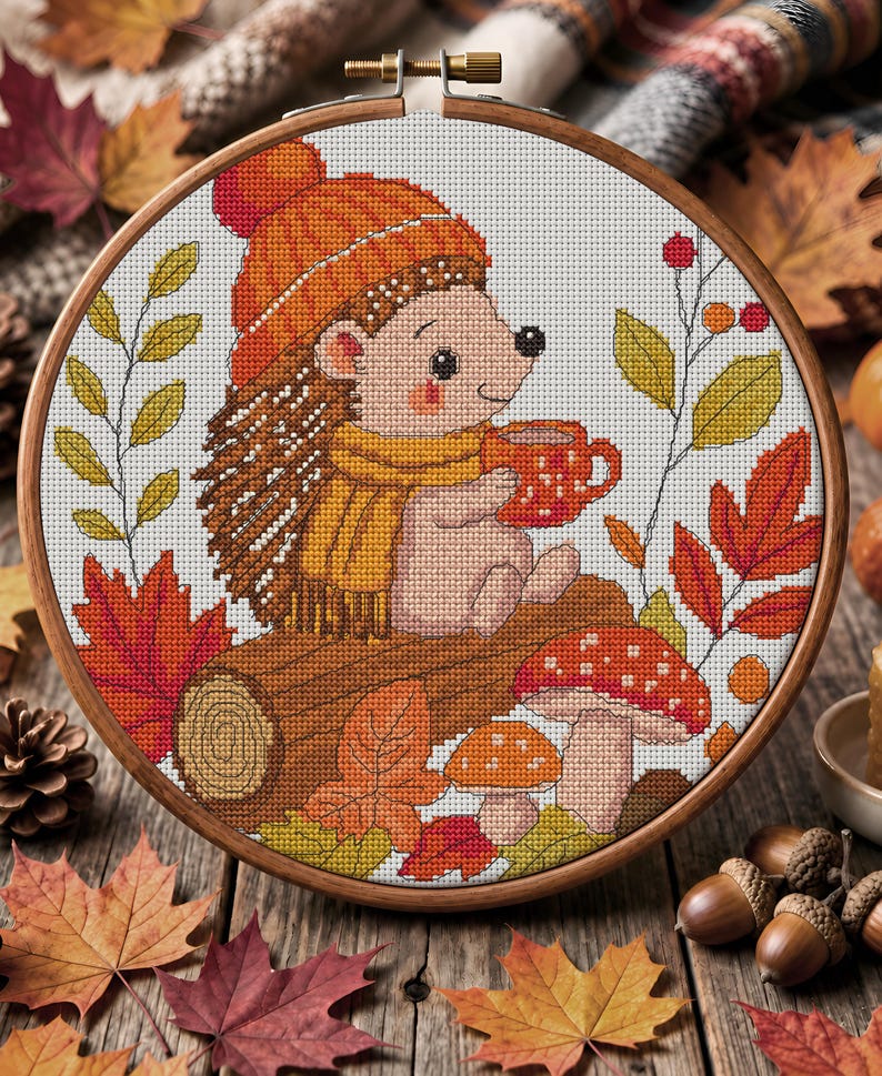May include: A circular wooden embroidery hoop containing a cross-stitch pattern of a hedgehog wearing an orange knit hat and a yellow scarf, holding a red mug. The scene includes a brown log, red and orange autumn leaves, and two mushrooms.