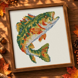 May include: A framed cross-stitch embroidery featuring a vibrant, multi-colored fish leaping upwards. The fish is rendered in shades of green, orange, yellow, and blue, set against a white Aida cloth background within a simple wooden frame.