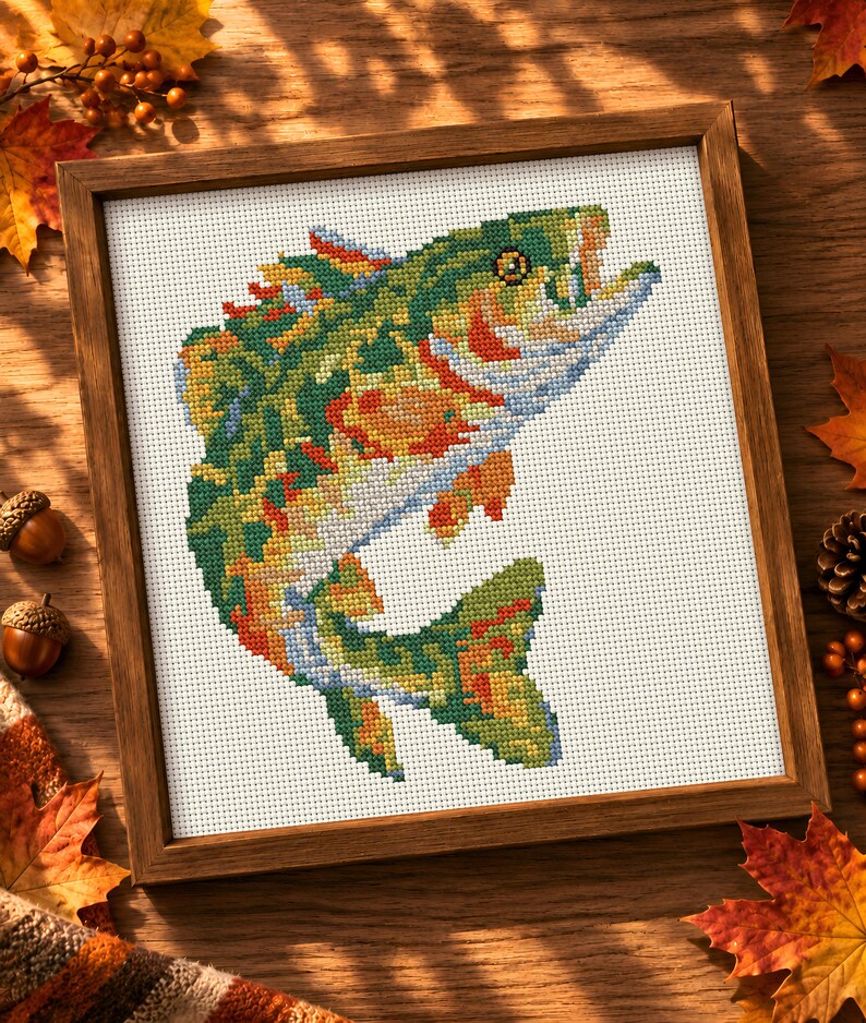 May include: A framed cross-stitch embroidery featuring a vibrant, multi-colored fish leaping upwards. The fish is rendered in shades of green, orange, yellow, and blue, set against a white Aida cloth background within a simple wooden frame.