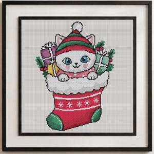 May include: A cross-stitch pattern featuring a white cat with blue eyes wearing a red and green striped hat, sitting inside a red Christmas stocking. The cat is surrounded by wrapped gift boxes, holly leaves, and berries.