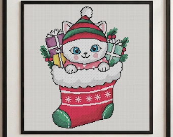 May include: A cross-stitch pattern featuring a white cat with blue eyes wearing a red and green striped hat, sitting inside a red Christmas stocking. The cat is surrounded by wrapped gift boxes, holly leaves, and berries.