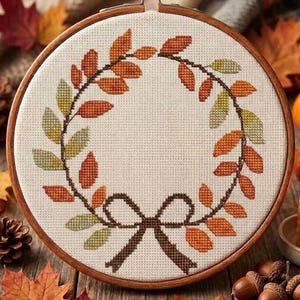 May include: A circular wooden embroidery hoop containing a cross-stitch pattern of an autumn leaf wreath. The design features orange, red, and green leaves arranged in a circle, tied at the bottom with a dark brown stitched bow on cream fabric.