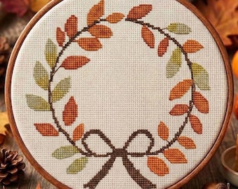 May include: A circular wooden embroidery hoop containing a cross-stitch pattern of an autumn leaf wreath. The design features orange, red, and green leaves arranged in a circle, tied at the bottom with a dark brown stitched bow on cream fabric.