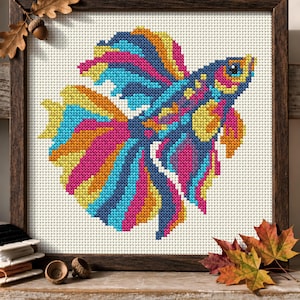 May include: A cross-stitch embroidery of a vibrant betta fish featuring bright pink, blue, and yellow threads on a white Aida cloth. The piece is encased in a dark brown wooden frame, showcasing intricate needlework detail and colourful patterns.