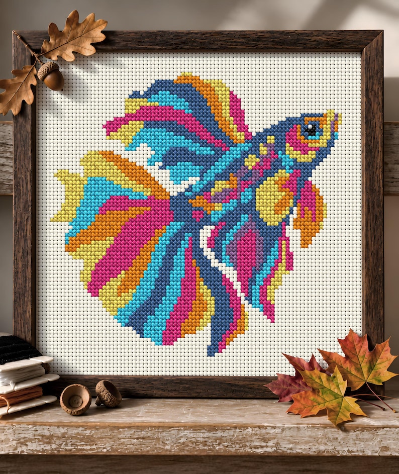 May include: A cross-stitch embroidery of a vibrant betta fish featuring bright pink, blue, and yellow threads on a white Aida cloth. The piece is encased in a dark brown wooden frame, showcasing intricate needlework detail and colourful patterns.