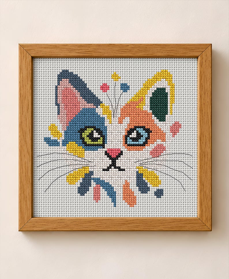 K&ouml;nnte beinhalten: Eine Kreuzstichstickerei eines Katzengesichts in einem hellen Holzrahmen. Die Katze hat eine wei&szlig;e Grundfarbe mit blauen, orangefarbenen, gelben und rosafarbenen Flecken. Sie hat gr&uuml;ne und blaue Augen, eine rosa Nase und schwarze Schnurrhaare.