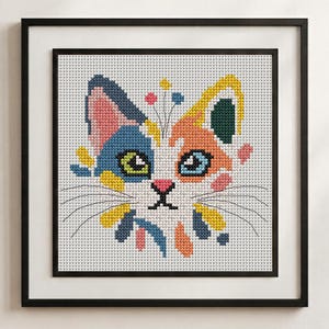 May include: A framed cross-stitch embroidery featuring a stylised cat head with a white base and patches of blue, orange, yellow, pink, and dark green. The cat has green and blue eyes, a pink nose, and black whiskers, all set against a white grid.
