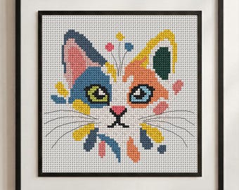 May include: A framed cross-stitch embroidery featuring a stylised cat head with a white base and patches of blue, orange, yellow, pink, and dark green. The cat has green and blue eyes, a pink nose, and black whiskers, all set against a white grid.