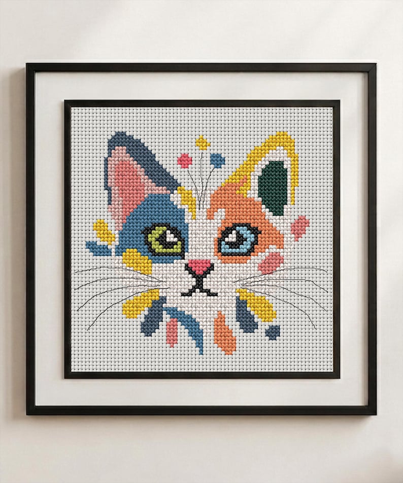 K&ouml;nnte beinhalten: Ein gerahmtes Kreuzstich-Stickbild, das einen stilisierten Katzenkopf auf wei&szlig;em Grund mit Flecken in Blau, Orange, Gelb, Rosa und Dunkelgr&uuml;n zeigt. Die Katze hat gr&uuml;ne und blaue Augen, eine rosa Nase und schwarze Schnurrhaare auf einem wei&szlig;en Gitter.