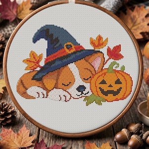 May include: A cross-stitch embroidery project in a wooden hoop featuring a sleeping puppy wearing a blue witch hat with an orange band. The puppy is positioned next to a carved orange jack-o'-lantern and several colourful autumn maple leaves.