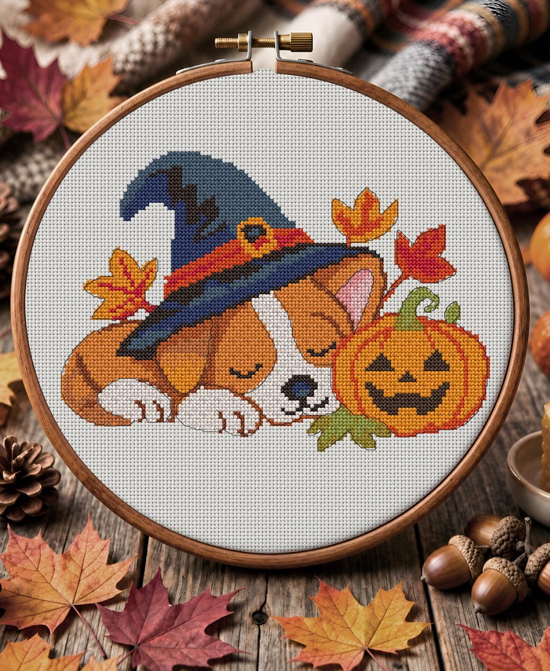 May include: A cross-stitch embroidery project in a wooden hoop featuring a sleeping puppy wearing a blue witch hat with an orange band. The puppy is positioned next to a carved orange jack-o'-lantern and several colourful autumn maple leaves.