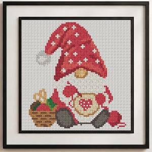 May include: A framed cross-stitch pattern featuring a gnome wearing a red hat with white snowflake accents. The gnome holds a tan mug with a red heart design, sits next to a brown basket of red apples, and has a ball of red yarn nearby.
