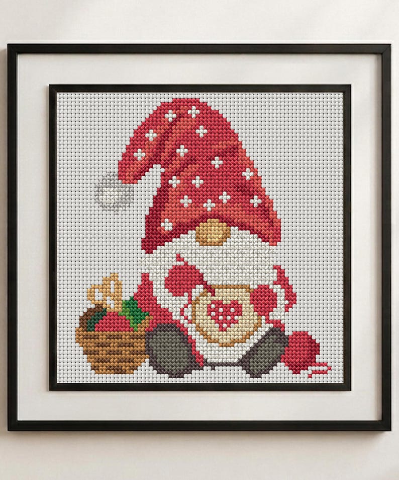 May include: A framed cross-stitch pattern featuring a gnome wearing a red hat with white snowflake accents. The gnome holds a tan mug with a red heart design, sits next to a brown basket of red apples, and has a ball of red yarn nearby.